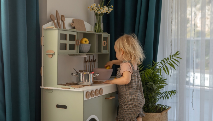 A young child smiles while playing with toy kitchen toys in a well-decorated, light-filled room. 